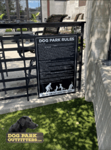 A Professional Rules & Wellness Sign - In-Ground Mount - Vertical Sign titled "Dog Park Rules" is mounted on a fence by an outdoor patio. Below, artificial grass displays a logo with a black dog and "Dog Park Outfitters" text.